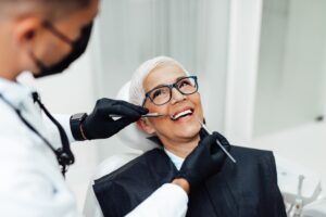 A woman having her veneers examined by a dentist. She is sitting back in the dentist's chair and smiling.